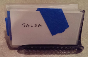 A business card holder with many cards in it. The front one says "salsa" and has blue painter's tape on it.